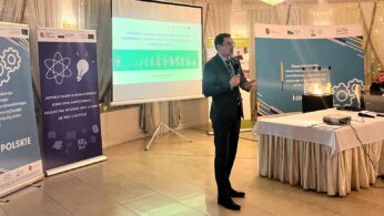 Mężczyzna w garniturze mówi do mikrofonu w dobrze oświetlonej sali konferencyjnej. Za nim znajduje się duży ekran projekcyjny wyświetlający zielony slajd prezentacji w języku polskim. Banery z logo i grafikami otaczają ekran, a na pierwszym planie widoczne są stoły publiczności.