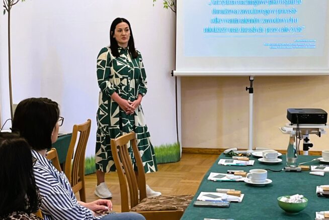 Kobieta w zielonej wzorzystej sukience stoi i przemawia obok wyświetlanej prezentacji. Kilka osób siedzi przy stole z filiżankami kawy, przekąskami i notatnikami. Na banerze widnieje logo Polski i UE, a na pierwszym planie znajduje się duży zielony stół.