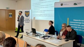 Mężczyzna stoi i mówi przez mikrofon w pobliżu ekranu projektora wyświetlającego polski tekst. Trzy osoby, dwie kobiety i jeden mężczyzna, siedzą przy stole z laptopami i papierami. Banery i niebieski wyświetlacz są widoczne za nimi w klasie.