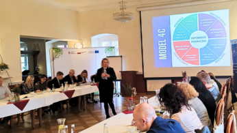 Kobieta stoi i przemawia do grupy siedzącej wokół stołów w kształcie litery U w sali konferencyjnej. Na dużym ekranie wyświetlany jest kolorowy diagram modelu 4C. Uczestnicy słuchają uważnie; notatki, napoje i jedzenie znajdują się na stołach w jasnej, eleganckiej przestrzeni.