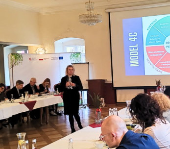 Kobieta stoi i przemawia do grupy siedzącej wokół stołów w kształcie litery U w sali konferencyjnej. Na dużym ekranie wyświetlany jest kolorowy diagram modelu 4C. Uczestnicy słuchają uważnie; notatki, napoje i jedzenie znajdują się na stołach w jasnej, eleganckiej przestrzeni.