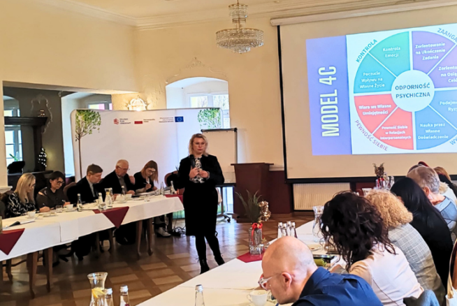 Kobieta stoi i przemawia do grupy siedzącej wokół stołów w kształcie litery U w sali konferencyjnej. Na dużym ekranie wyświetlany jest kolorowy diagram modelu 4C. Uczestnicy słuchają uważnie; notatki, napoje i jedzenie znajdują się na stołach w jasnej, eleganckiej przestrzeni.