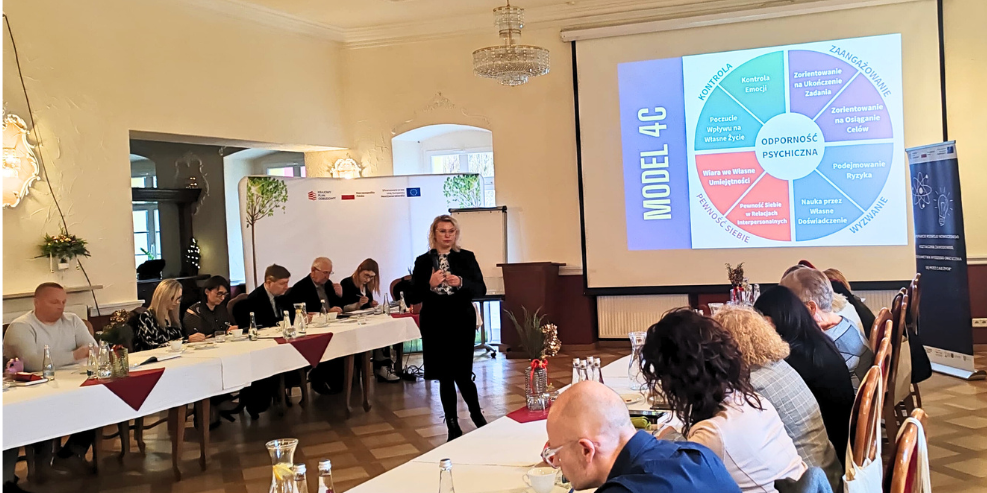 Kobieta stoi i przemawia do grupy siedzącej wokół stołów w kształcie litery U w sali konferencyjnej. Na dużym ekranie wyświetlany jest kolorowy diagram modelu 4C. Uczestnicy słuchają uważnie; notatki, napoje i jedzenie znajdują się na stołach w jasnej, eleganckiej przestrzeni.