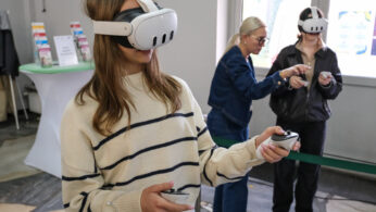 Dwie osoby w swetrach w paski noszą gogle VR i trzymają kontrolery, zanurzone w wirtualnym doświadczeniu. W tle starsza kobieta gestykuluje, pomagając innej osobie trzymającej urządzenie. Znajdują się w jasnym pomieszczeniu z materiałami informacyjnymi w pobliżu.