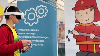 Młody chłopiec noszący zestaw VR i trzymający kontrolery stoi w pobliżu banera z polskim tekstem i dużą ilustracją uśmiechniętego strażaka trzymającego wąż. Scena wygląda na wydarzenie edukacyjne lub szkoleniowe.