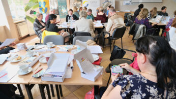 Grupa dorosłych siedzi przy stołach w jasno oświetlonym pokoju, zaangażowana w dyskusję i papierkową robotę. Na stołach leżą filiżanki kawy, ciastka i notatniki. W tle widoczne są plakaty i banery z logo i grafiką.