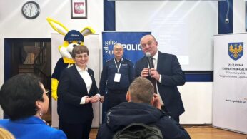 Grupa osób, w tym dwóch policjantów, kobieta w garniturze i mężczyzna z mikrofonem, stoją w pomieszczeniu. Za nimi znajduje się osoba w żółtym kostiumie maskotki policyjnej i oznakowanie policyjne. Członkowie publiczności są widoczni na pierwszym planie.
