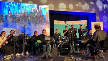 Zespół występuje na scenie z różnymi instrumentami, w tym perkusją, keyboardem, gitarą elektryczną i instrumentami dętymi. Kilku muzyków siedzi, podczas gdy inni stoją. Dyrygent stoi przed grupą, a duży ekran wyświetla ich obraz na żywo w tle.