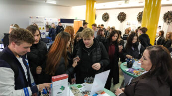 Grupa studentów gromadzi się wokół stołu z broszurami i długopisami na imprezie wewnętrznej. Kobieta w garniturze stoi w pobliżu, prawdopodobnie pomagając. W tle widać więcej osób i plakaty informacyjne. Pokój ma żółte filary i jasne oświetlenie.