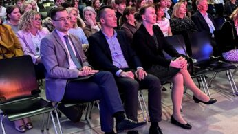 Duża grupa ludzi siedzi uważnie w rzędach krzeseł na imprezie wewnętrznej. Na pierwszym planie trzy osoby siedzą w pierwszym rzędzie, ubrane w stroje biznesowe, skupione na scenie. W tle widać więcej siedzących uczestników w przestronnej, dobrze oświetlonej sali.