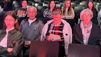 Grupa ludzi siedzi w rzędach czarnych krzeseł podczas imprezy w pomieszczeniu. Cztery starsze osoby i młoda kobieta siedzą w pierwszym rzędzie, przodem do kamery. Kolejni uczestnicy, w różnym wieku, siedzą za nimi, a w tle widoczna jest scena lub ekran.