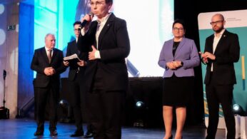 Pięć osób stoi na scenie w formalnych strojach. Kobieta z przodu mówi do mikrofonu. Cztery inne osoby - trzech mężczyzn i jedna kobieta - stoją za nią, trzymając dokumenty lub stojąc ze splecionymi rękami. Jasne światła i duży ekran tworzą nowoczesne, profesjonalne otoczenie.