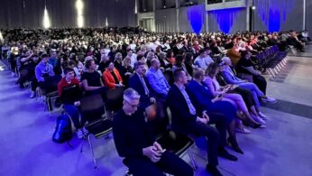 Duża publiczność siedzi w rzędach krzeseł w przestronnej, nowoczesnej sali z wysokimi sufitami i zasłonami oświetlonymi niebieskim światłem. Większość osób zwrócona jest twarzami do przodu, niektórzy używają telefonów, a miejsca po jednej stronie są w większości puste. Atmosfera jest skupiona i wyczekująca.
