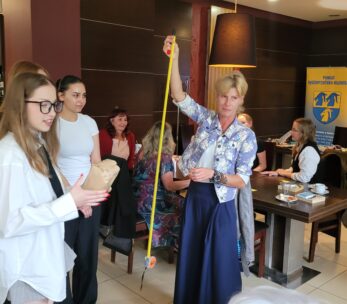 Kobieta trzyma żółtą taśmę mierniczą w kawiarni lub sali konferencyjnej, na co patrzy kilka kobiet. Ludzie siedzą przy stolikach z papierami i napojami. Banery i lampy są widoczne w tle, nadając profesjonalną, ale swobodną atmosferę.