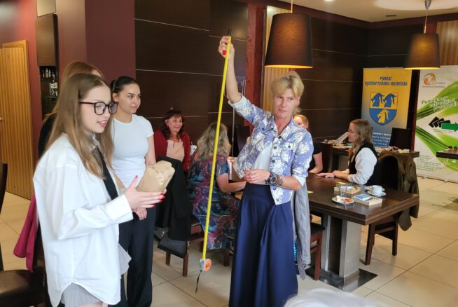 Kobieta trzyma żółtą taśmę mierniczą w kawiarni lub sali konferencyjnej, na co patrzy kilka kobiet. Ludzie siedzą przy stolikach z papierami i napojami. Banery i lampy są widoczne w tle, nadając profesjonalną, ale swobodną atmosferę.
