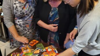 Trzy kobiety stoją wokół stołu pokrytego kolorowymi ilustrowanymi kartami. Przyglądają się sobie uważnie i wskazują na karty, wydając się je omawiać. Sceneria jest wewnątrz, a inne osoby są rozmyte w tle.