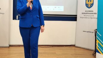 Kobieta w jasnoniebieskim garniturze mówi do mikrofonu, stojąc na drewnianej podłodze. Za nią znajdują się banery z polskim tekstem i oficjalne logo, w tym żółty herb z orłem, a w tle ekran projekcyjny wyświetlający prezentację.