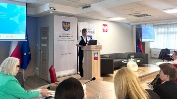 Kobieta stoi na podium, prowadząc prezentację w sali konferencyjnej. Uczestnicy siedzą i słuchają. Za prelegentką znajdują się banery z herbami, polska flaga i ekran wyświetlający slajd z tekstem. Pokój ma nowoczesny wystrój i duże okna z żaluzjami.