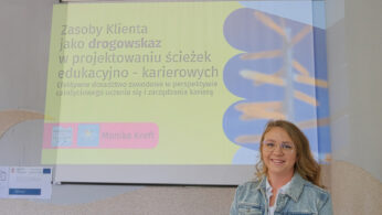 Kobieta w okularach i blond włosach, ubrana w jasnoniebieską marynarkę i białą koszulę, stoi uśmiechnięta przed wyświetlanym na ścianie slajdem prezentacji w języku polskim. Slajd omawia zasoby klienta w planowaniu kariery i ścieżki edukacyjnej.