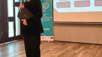 Mężczyzna w garniturze trzyma mikrofon i teczkę, przemawiając podczas wydarzenia w pomieszczeniu. Za nim znajdują się banery i duży ekran wyświetlający slajd zatytułowany 