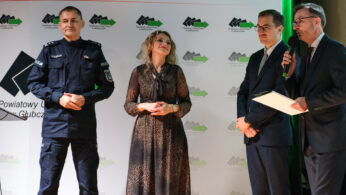 Cztery osoby stoją na scenie przed banerem z polskim tekstem. Od lewej: mężczyzna w policyjnym mundurze, uśmiechnięta kobieta w brązowej sukience i dwóch mężczyzn w garniturach - jeden trzyma mikrofon i papier, najwyraźniej przemawiając. Atmosfera jest formalna.