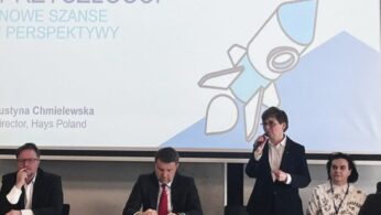 Wydarzenie panelowe Hays Poland. Justyna Chmielewska, Dyrektor w Hays Poland, stoi i mówi do mikrofonu. Trzech panelistów siedzi przy stole. Na slajdzie prezentacji widnieje napis Kompetencje Przyszłości - Nowe Szanse i Perspektywy z grafiką rakiety.