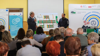 Mężczyzna w garniturze przemawia do mikrofonu obok umundurowanego funkcjonariusza policji przed siedzącą publicznością. Za nimi znajduje się duża tablica z diagramami i banerami wyświetlającymi logo, tekst i grafikę związaną z doradztwem zawodowym i samorządem lokalnym.