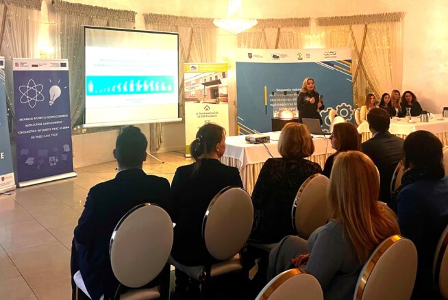 Kobieta stoi i przemawia na podium w sali konferencyjnej, zwracając się do publiczności siedzącej w rzędach. Slajdy prezentacji są wyświetlane na ekranie, a banery informacyjne są wyświetlane po obu stronach. Kilka osób siedzi przy stole obok prelegentki.