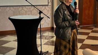 Kobieta o krótkich blond włosach, w okularach i skórzanej kurtce stoi trzymając mikrofon w pokoju z podłogą w kratkę. Za nią znajduje się ekran projektora wyświetlający polską prezentację zatytułowaną By zawód nie sprawił zawodu.