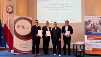 Cztery osoby stoją w pomieszczeniu, uśmiechają się i trzymają dokumenty. Za nimi znajduje się ekran wyświetlający polski tekst o spotkaniu inauguracyjnym 29.10.2024 r. w Warszawie, banery z logo ORE i FRSE oraz flagi. Pokój ma beżowe ściany i niebieski dywan.
