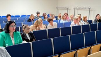 Grupa kobiet siedzi w klasie lub sali wykładowej z niebieskimi krzesłami, uważnie słuchając. Niektóre mają notatniki i długopisy. Pomieszczenie jest jasne, z wieszakami z tyłu i w większości pustymi rzędami na pierwszym planie.