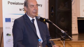 Mężczyzna w ciemnym garniturze i krawacie przemawia na drewnianym podium z mikrofonami. Na podium stoi butelka z wodą. Za nim znajdują się banery z polskim tekstem, logo i flagą UE. W tle widoczne są duże głośniki i scena z ekranem.