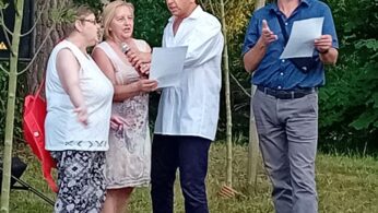 Cztery dorosłe osoby stoją na zewnątrz na trawie, otoczone młodymi drzewami. Jedna kobieta trzyma mikrofon, inna kobieta i mężczyzna czytają z kartki, podczas gdy inny mężczyzna w niebieskiej koszuli i okularach gestykuluje i trzyma kartkę. Grupa wydaje się być zaangażowana w dyskusję lub przedstawienie.