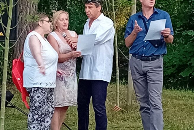 Cztery dorosłe osoby stoją na zewnątrz na trawie, otoczone młodymi drzewami. Jedna kobieta trzyma mikrofon, inna kobieta i mężczyzna czytają z kartki, podczas gdy inny mężczyzna w niebieskiej koszuli i okularach gestykuluje i trzyma kartkę. Grupa wydaje się być zaangażowana w dyskusję lub przedstawienie.