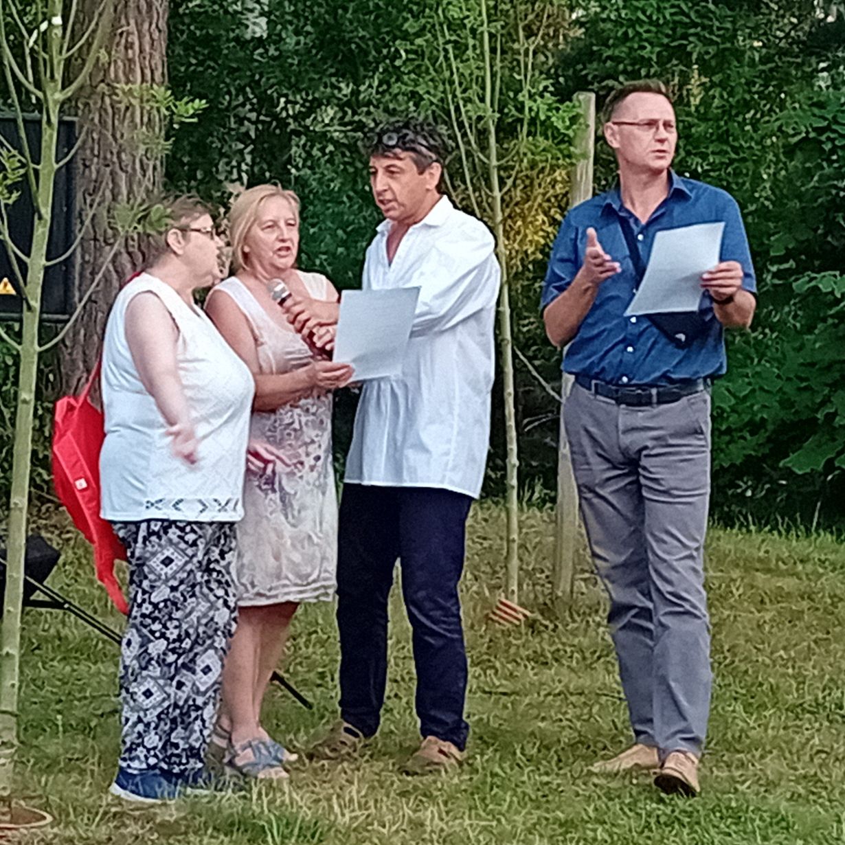 Cztery dorosłe osoby stoją na zewnątrz na trawie, otoczone młodymi drzewami. Jedna kobieta trzyma mikrofon, inna kobieta i mężczyzna czytają z kartki, podczas gdy inny mężczyzna w niebieskiej koszuli i okularach gestykuluje i trzyma kartkę. Grupa wydaje się być zaangażowana w dyskusję lub przedstawienie.