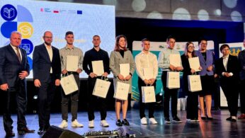Grupa 11 osób stoi na scenie, większość z nich trzyma torby z prezentami i certyfikatami. Ubrani są w stroje biznesowe i eleganckie. Za nimi duży ekran i banery wyświetlające logo i tekst w języku polskim. Wygląda to na ceremonię wręczenia nagród.