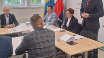Pięć osób znajduje się w sali konferencyjnej z dużymi oknami i flagami, w tym Polski. Trzy osoby siedzą przy stole naprzeciwko dwóch innych; jedna ze stojących osób przemawia. Na stole leżą papiery i butelki, a nastrój wydaje się formalny i profesjonalny.