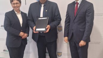 Trzy osoby w formalnych strojach stoją w pomieszczeniu. Mężczyzna w centrum trzyma oprawiony certyfikat i uśmiecha się, a po jego lewej stronie stoi kobieta, a po prawej mężczyzna. Za nimi znajduje się baner z polskim tekstem i regionalnymi emblematami.
