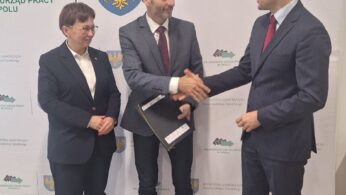 Trzy osoby w strojach biznesowych stoją w pomieszczeniu na tle oficjalnych logo i tekstu. Dwóch mężczyzn w garniturach podaje sobie ręce; jeden trzyma teczkę. Kobieta w garniturze stoi obok nich, uśmiechając się. Otoczenie wydaje się formalne, prawdopodobnie ceremonia lub oficjalne wydarzenie.