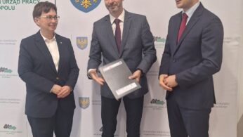 Trzy profesjonalnie ubrane osoby stoją i uśmiechają się przed markowym tłem z herbami i polskim tekstem. Osoba w środku trzyma teczkę z dokumentami. Wszyscy wyglądają na zaangażowanych i mają na sobie stroje biznesowe, dwóch w garniturach, a jeden w marynarce i spodniach.