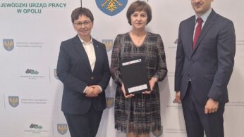 Trzy dorosłe osoby stoją w pomieszczeniu przed banerami z polskim tekstem i emblematami. Kobieta w centrum trzyma czarną teczkę. Wszyscy troje są formalnie ubrani, uśmiechnięci i zwróceni w stronę kamery. Tło sugeruje oficjalne lub profesjonalne wydarzenie.
