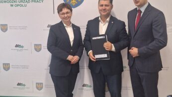 Trzy osoby w formalnych strojach stoją uśmiechnięte przed tłem z polskim tekstem i logo. Mężczyzna w centrum trzyma dokument. Tło zawiera herby i znaki Wojewódzkiego Urzędu Pracy w Opolu i Jednostki Samorządu Województwa Opolskiego.