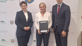 Trzy osoby stoją w pomieszczeniu na jasnej podłodze przed banerem z oficjalnym logo i polskim tekstem. Kobieta w centrum trzyma czarną teczkę; wszyscy są ubrani formalnie i uśmiechają się do kamery.