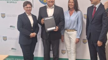 Cztery osoby stoją w pomieszczeniu, uśmiechając się do kamery przed banerami Wojewódzkiego Urzędu Pracy w Opolu. Osoba w centrum trzyma czarną teczkę. Wszyscy ubrani są w stroje służbowe; dwie kobiety i dwóch mężczyzn pozuje razem.