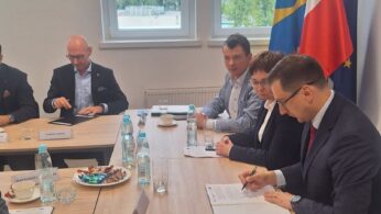 Kilka osób w formalnych strojach siedzi przy stole konferencyjnym z dokumentami, butelkami wody i przekąskami. Dwóch mężczyzn na pierwszym planie podpisuje dokumenty. Za nimi znajdują się okna i widoczne są flagi Polski, Unii Europejskiej i regionalne.