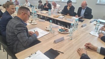 Grupa ludzi w strojach biznesowych siedzi wokół dużego stołu konferencyjnego z dokumentami, butelkami wody, ciastkami i słodyczami. Niektórzy piszą lub czytają dokumenty, inni uważnie słuchają. Na stole znajdują się tabliczki z nazwiskami, a przez okna wpada jasne, naturalne światło.