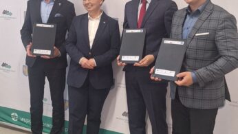 Cztery osoby stoją obok siebie, uśmiechając się, przed tłem z logo i polskim tekstem. Trzech mężczyzn w garniturach trzyma dokumenty, a obok nich stoi kobieta w ciemnym garniturze. Scena wygląda na formalną, prawdopodobnie podczas ceremonii podpisania lub wręczenia nagród.