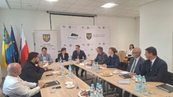 Grupa dziesięciu osób w strojach biznesowych siedzi wokół stołu konferencyjnego z butelkami wody, przekąskami i papierami. Za nimi znajdują się flagi, banery i logo z żółtym orłem na niebieskim tle. Spotkanie odbywa się w nowoczesnym, jasnym pomieszczeniu biurowym.