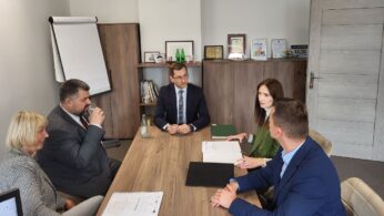 Pięć profesjonalnie ubranych osób siedzi przy drewnianym biurowym stole i dyskutuje. Na stole leżą papiery i foldery. Za nimi znajdują się półki z nagrodami, książkami i flipchartem. Biuro ma schludny, formalny wygląd.