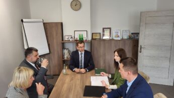 Pięć osób w strojach biznesowych siedzi przy drewnianym stole konferencyjnym w nowoczesnym biurze. Na stole leżą papiery i foldery. W tle zegar, półka z nagrodami i zamknięte drzwi. Wszyscy wydają się być zaangażowani w dyskusję.